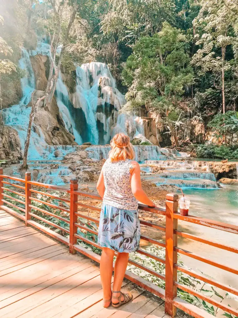 Em stands at a wooden railing sipping an iced drink while admiring the turquoise tiers of Kuang Si Falls, framed by jungle foliage and morning light.