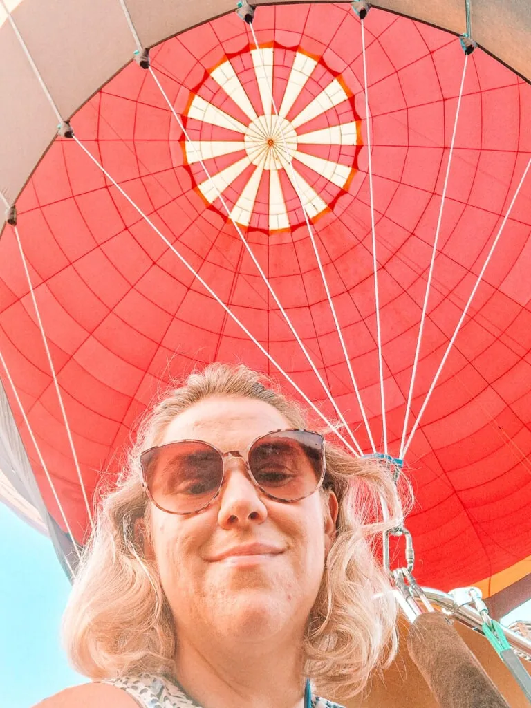Em takes a selfie from a hot air balloon basket, smiling under the vibrant red canopy with white spokes radiating out from the centre.