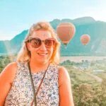 Em smiles with hot air balloons rising behind her over a rural landscape of fields, trees and mountains during a sunrise ride in Laos, part of her 3 week Laos itinerary.