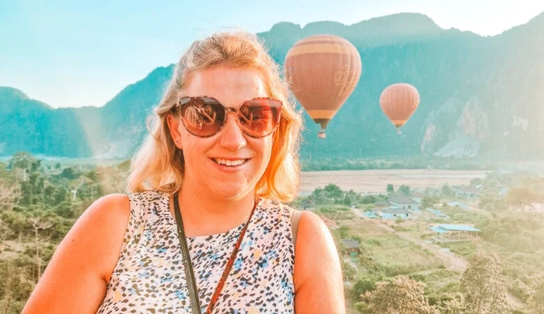 Em smiles with hot air balloons rising behind her over a rural landscape of fields, trees and mountains during a sunrise ride in Laos, part of her 3 week Laos itinerary.