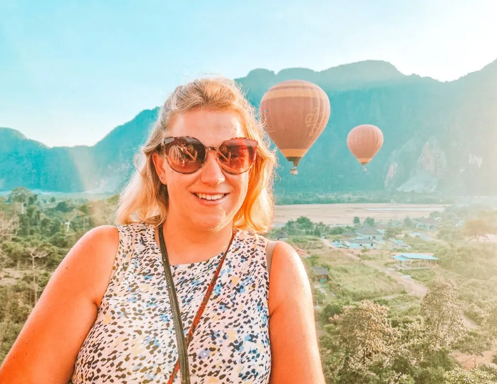 Em smiles in the foreground wearing sunglasses and a sleeveless top, with hot air balloons floating behind her over the lush mountains and rice fields of Vang Vieng.