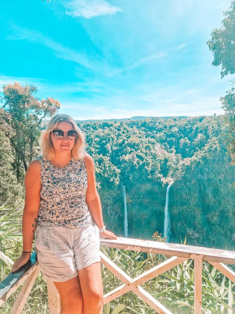 Em leans casually against a white wooden railing at a viewpoint overlooking twin waterfalls pouring into a jungle valley, a highlight of her 3 week Laos itinerary.