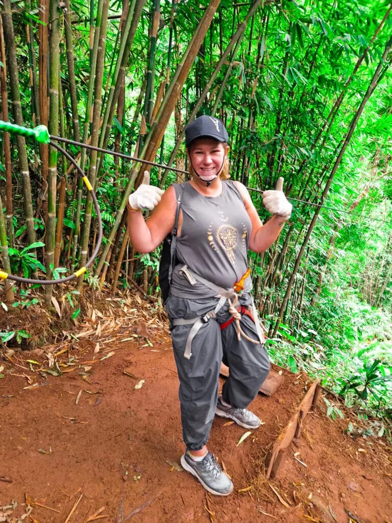 Em stands on a muddy trail in the jungle wearing a helmet, gloves and harness, giving two thumbs up before a ziplining adventure through a bamboo forest.