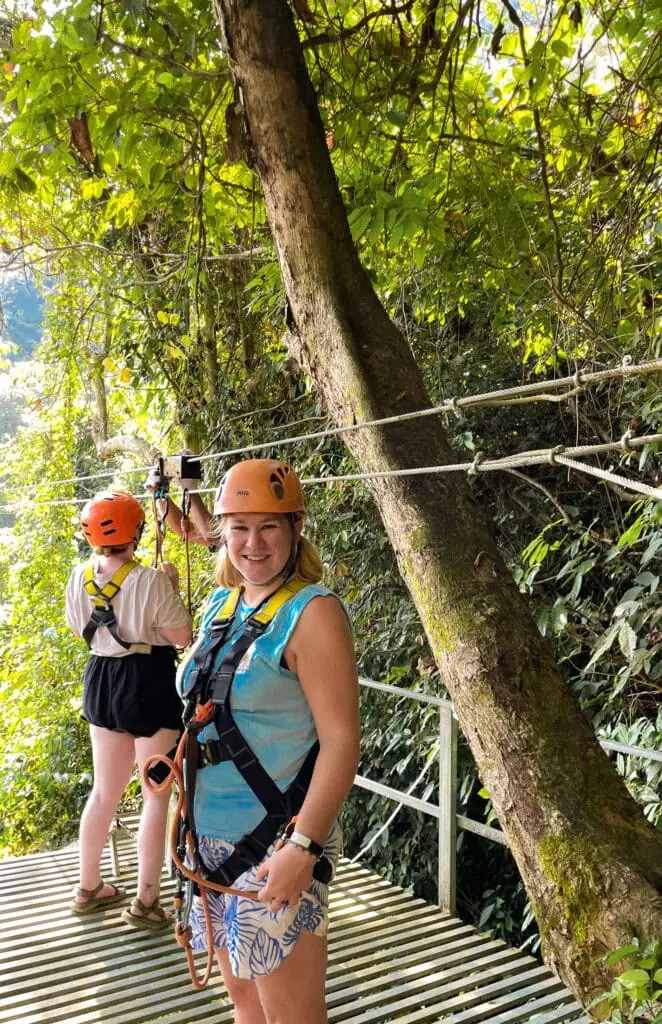 Wearing a helmet and harness, Em smiles confidently on a forest platform before ziplining through the trees as part of an eco-adventure in Laos.