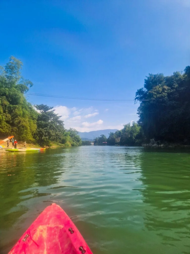 The front of a red kayak floats on a calm green river with forested banks and distant mountains under a sunny sky, a peaceful moment during the 2 week South to North Laos itinerary.
