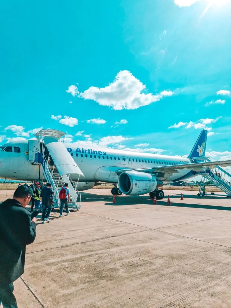 Passengers board a Lao Airlines plane via stairs on the tarmac under a vivid blue sky, with scattered clouds and a wide concrete runway.