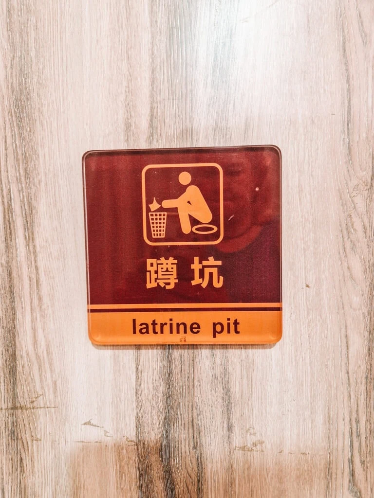 A toilet sign with Chinese and English text reads "latrine pit" and shows a graphic of a person squatting and disposing of tissue in a bin, fixed to a wooden wall.