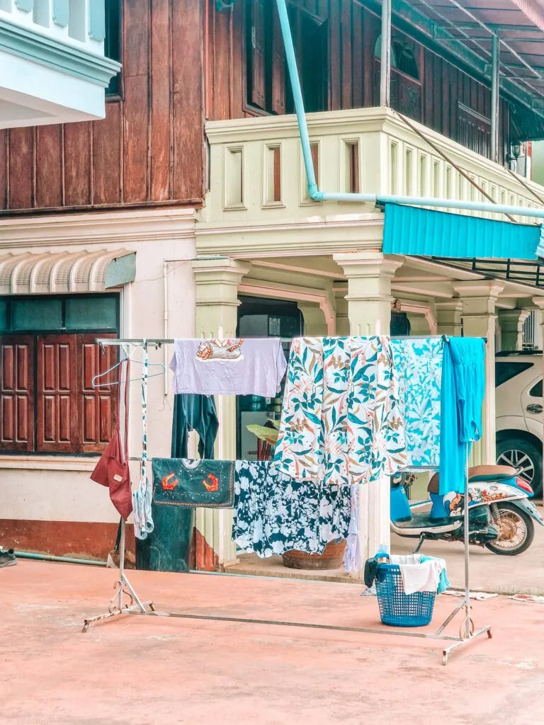 A metal rack full of colourful laundry, including floral prints and a crab-themed towel, stands outside a traditional Lao house with wooden and pastel-green walls.