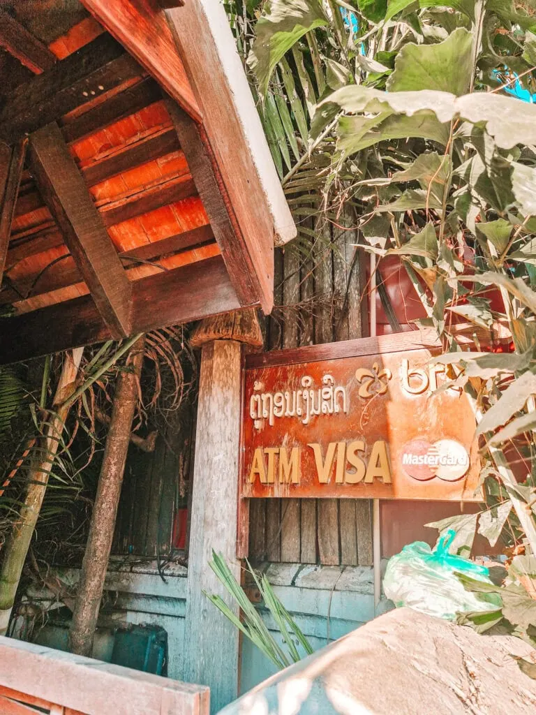 A faded wooden ATM sign with VISA and Mastercard logos is partially hidden by overgrown tropical plants under a traditional wooden awning in Laos.
