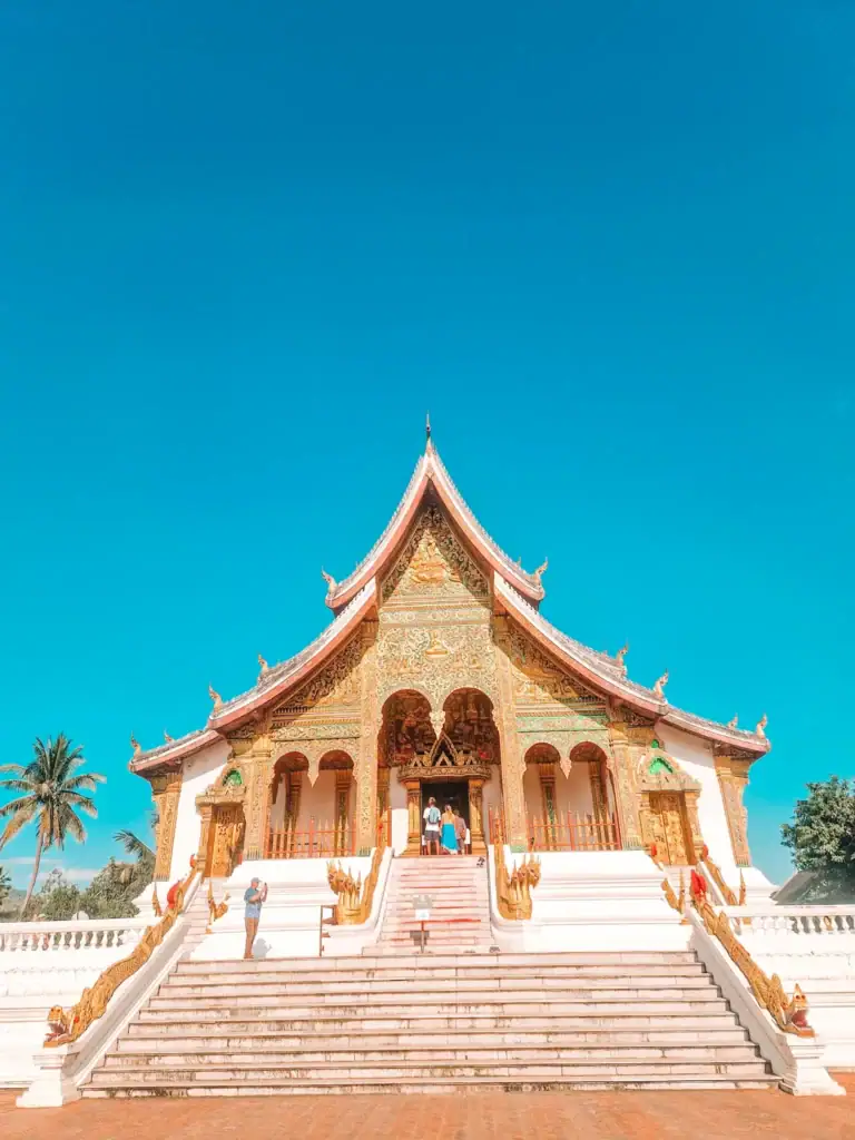 The ornate entrance to Haw Pha Bang temple in Luang Prabang stands tall under a clear blue sky, with golden carvings, red and green detailing, and a grand staircase welcoming visitors.