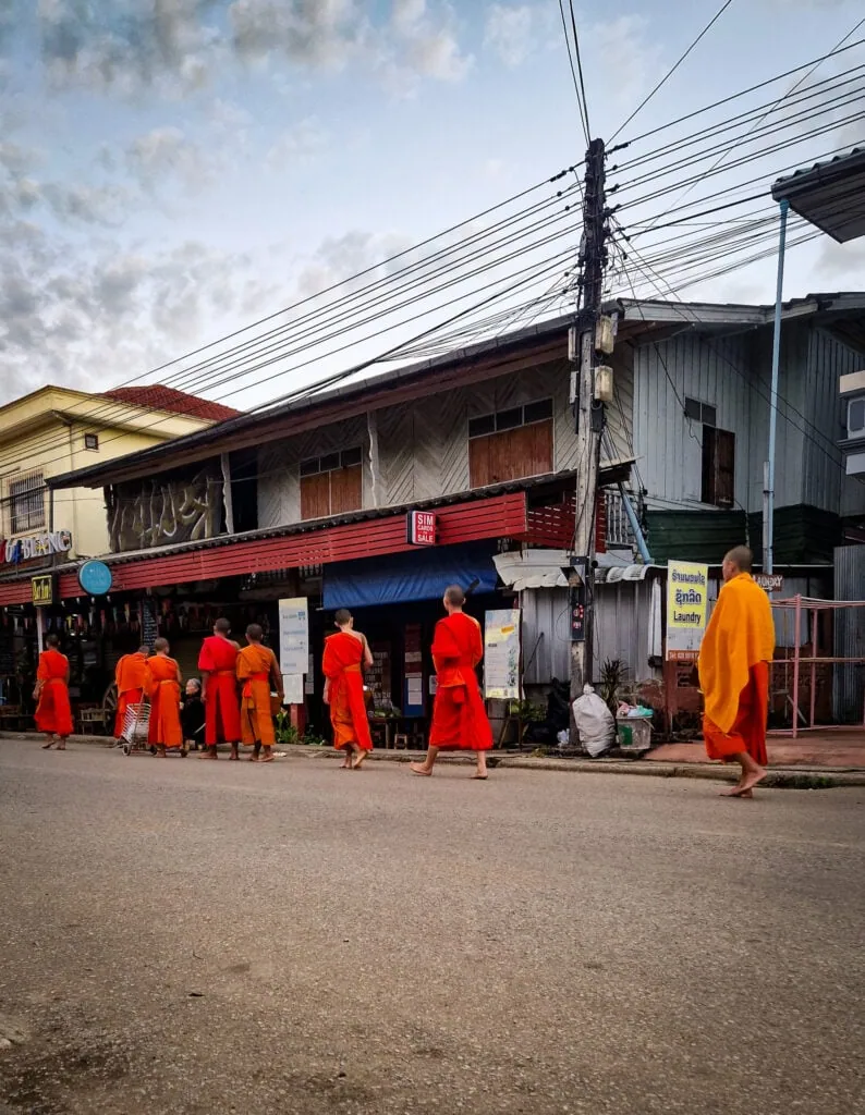 A group of young Buddhist monks in saffron robes walk barefoot in a line through a quiet town at dawn during the traditional alms-giving ceremony.