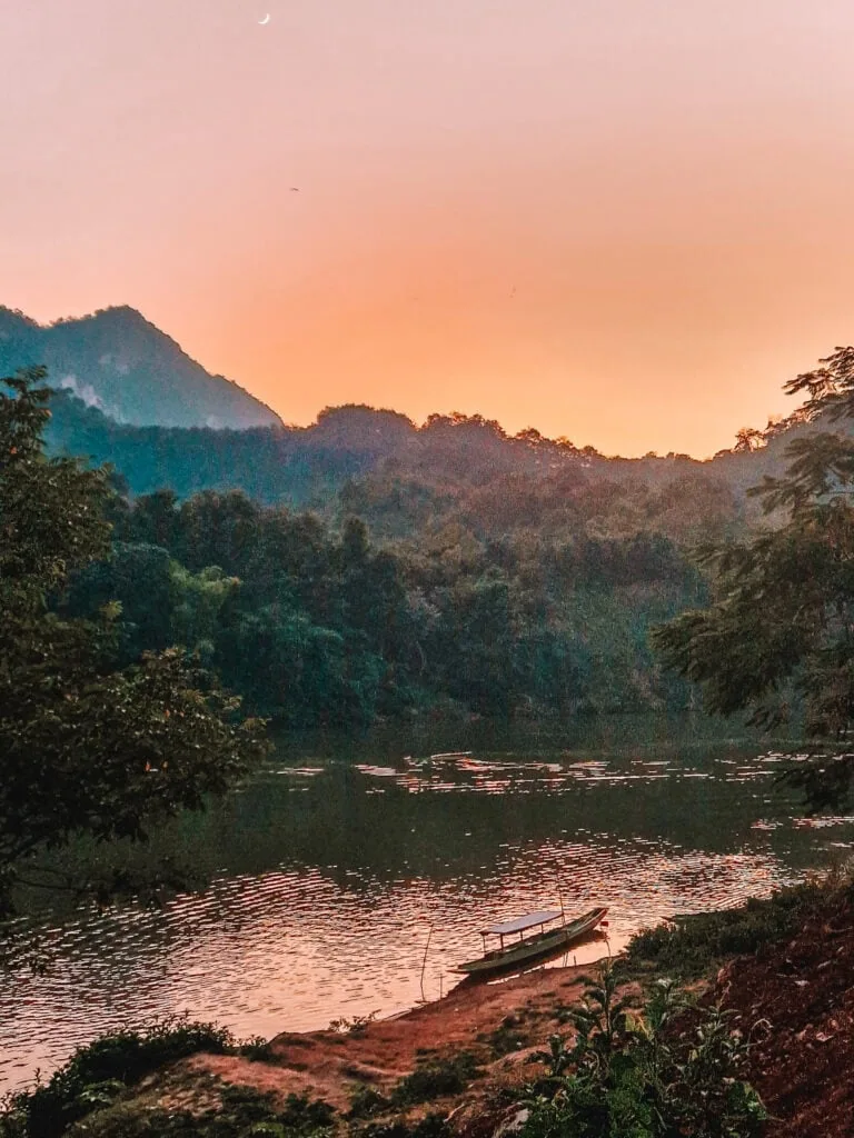 A small wooden boat rests on a still riverbank at dusk, backed by shadowy mountains and a glowing orange sky. Peaceful evenings like this are part of the magic of a 2 week north Laos itinerary.