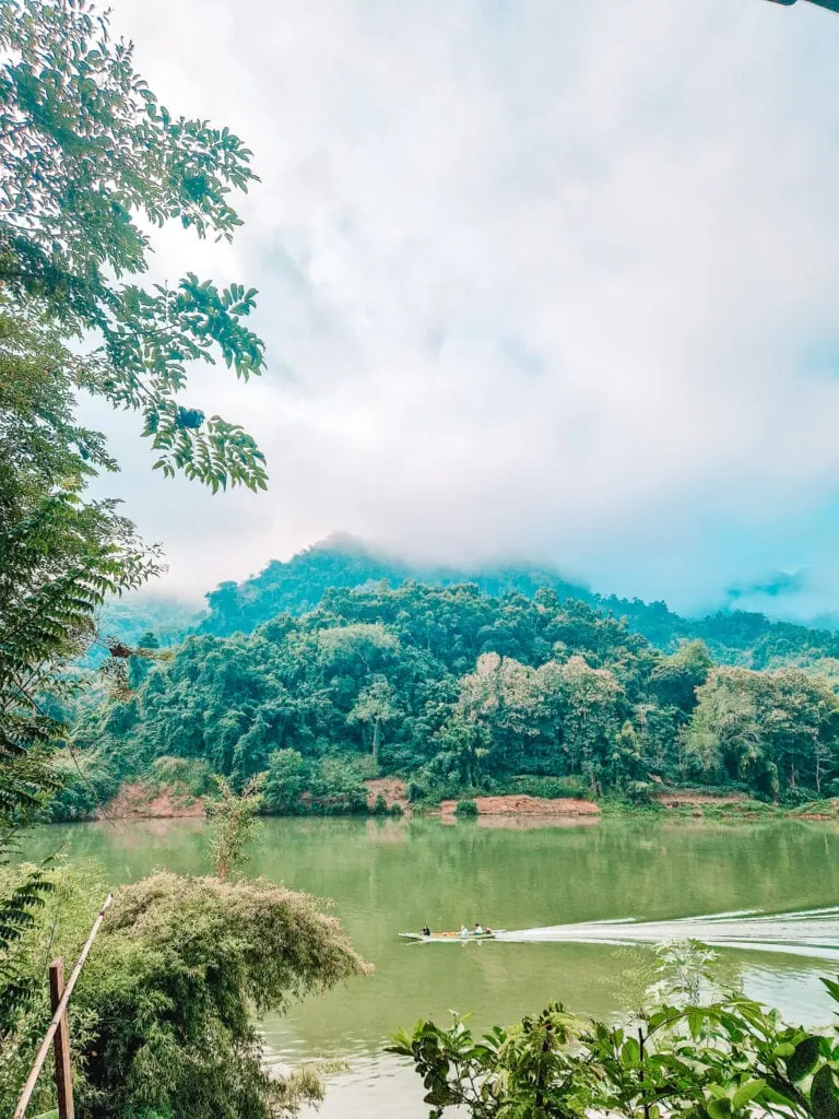 A misty morning view of a calm river with a small motorboat carrying passengers, framed by thick jungle and rolling green hills. Remote riverside moments like this define a 2 week north Laos itinerary.