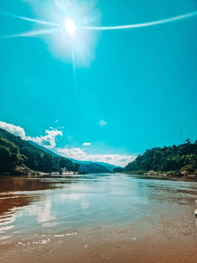 The Nam Ou River glistens in the midday sun, winding between forested hills and under a vivid blue sky in rural Laos.