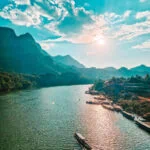 A longtail boat cruises along the Nam Ou River at sunset with golden light reflecting on the water and mountains in the distance. One of the most scenic views during a 2 week north Laos itinerary.
