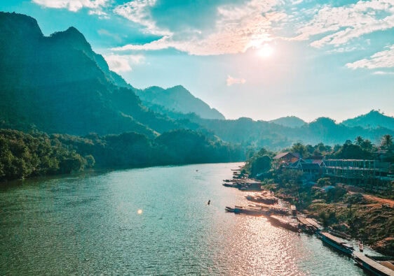 A longtail boat cruises along the Nam Ou River at sunset with golden light reflecting on the water and mountains in the distance. One of the most scenic views during a 2 week north Laos itinerary.