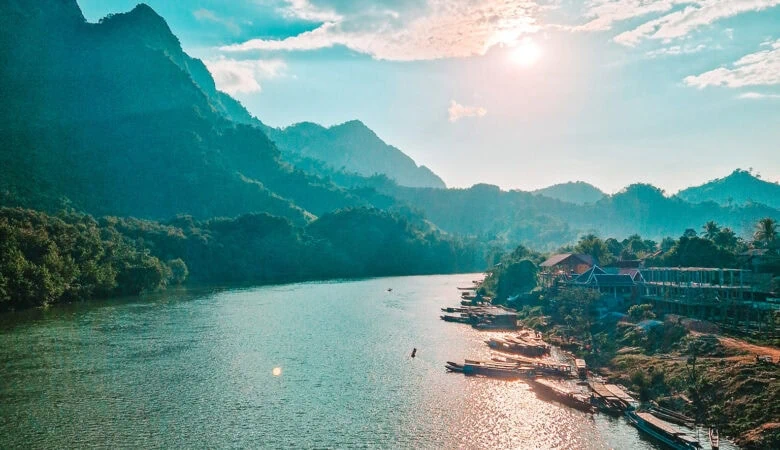 A longtail boat cruises along the Nam Ou River at sunset with golden light reflecting on the water and mountains in the distance. One of the most scenic views during a 2 week north Laos itinerary.