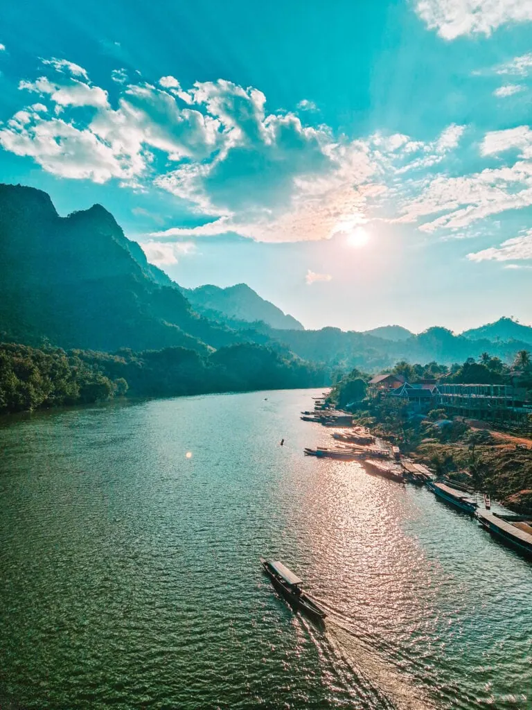 A longtail boat glides along the Nam Ou River as the sun sets over misty mountains and riverside buildings in northern Laos.