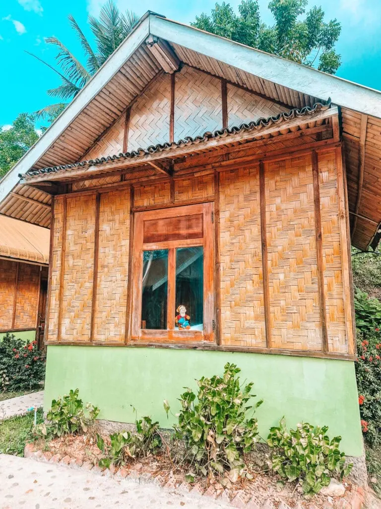 A woven bamboo bungalow with a green base and tin roof stands in a small garden, with a colourful soft toy visible on the windowsill.