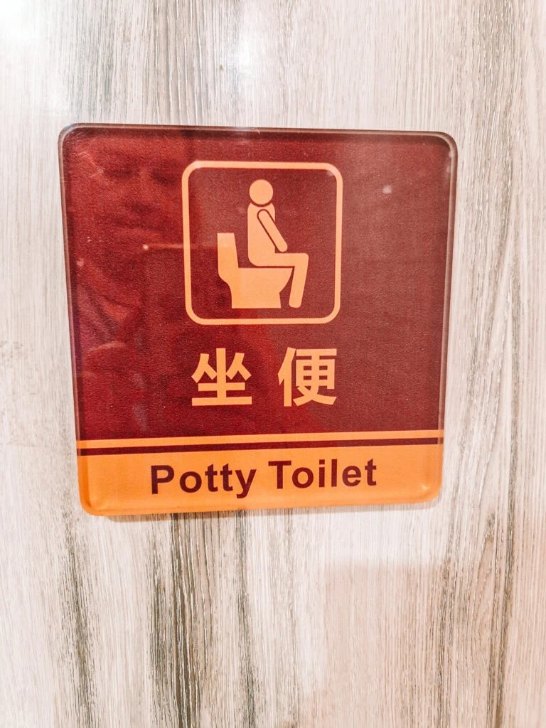 A toilet sign on wood-patterned wall shows a person sitting on a Western-style toilet, labelled “Potty Toilet” in English and Chinese characters.