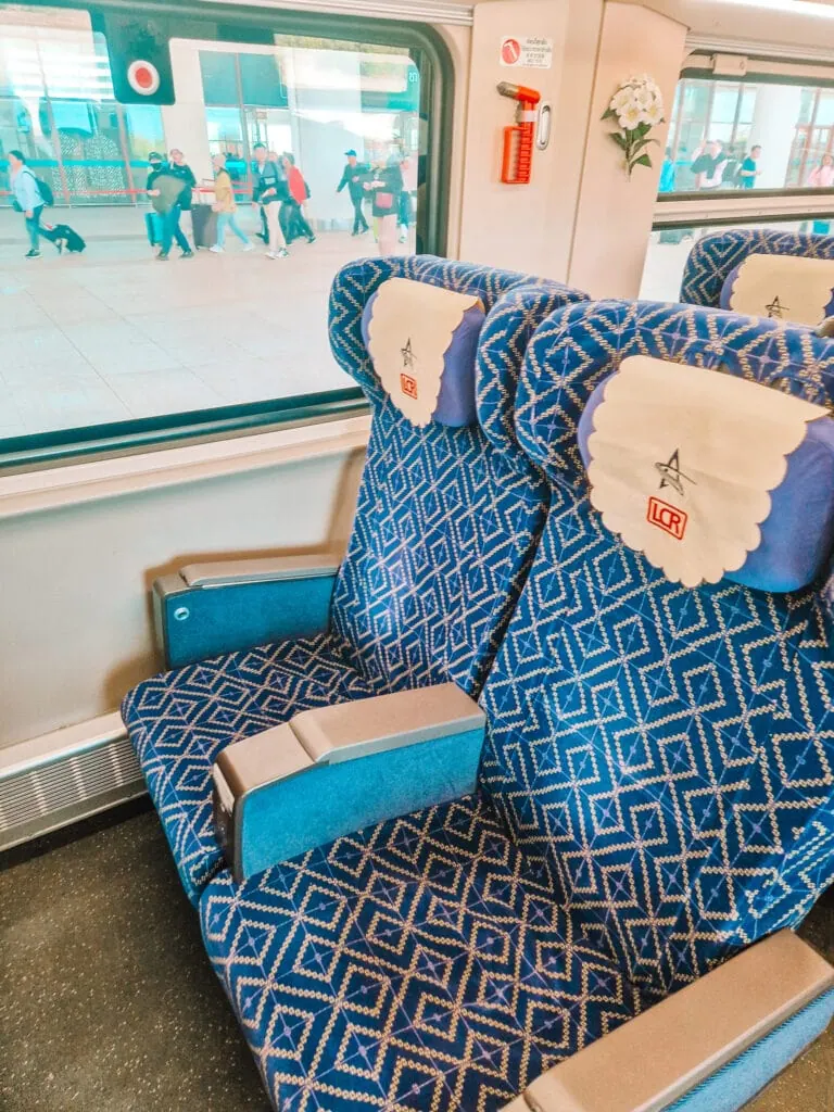 Two patterned blue train seats with headrest covers marked “LCR” face the window of a modern Lao railway carriage, with passengers visible outside on the platform.