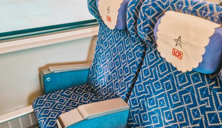 Two patterned blue train seats with headrest covers marked “LCR” face the window of a modern Lao railway carriage, with passengers visible outside on the platform.
