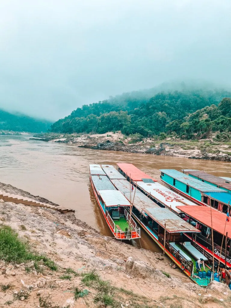 Several long, colourful slow boats are moored on the muddy banks of the Mekong River, with mist-covered jungle hills rising in the background.