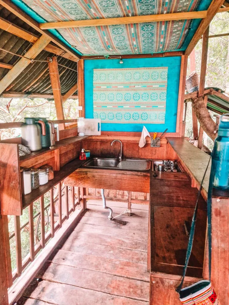 A rustic wooden kitchen built into a treehouse features basic cooking essentials, patterned teal walls and an open-air layout nestled in the jungle.