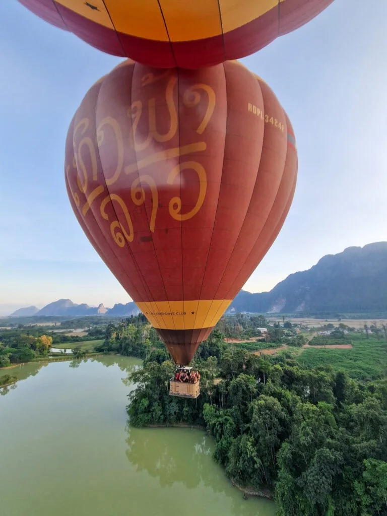 A red and yellow hot air balloon with Lao script floats above green countryside and a river, offering scenic views from the sky.