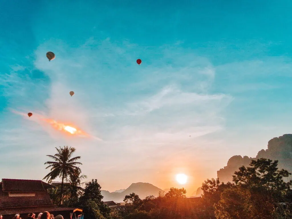 Hot air balloons rise above the rooftops and palm trees of Vang Vieng at sunrise, with the sun casting a soft glow over the limestone mountains.
