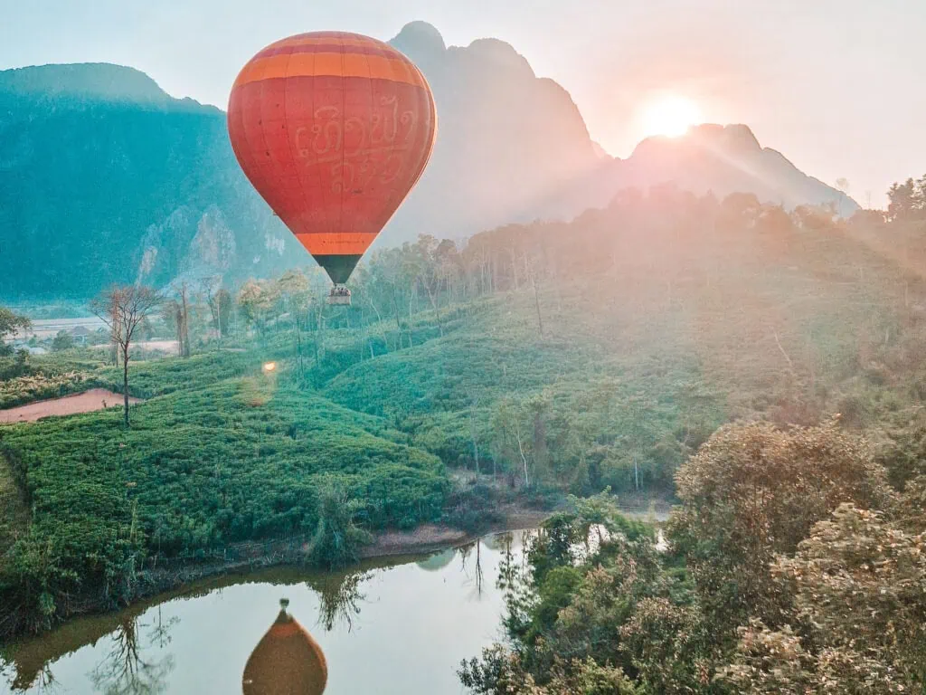 A red and orange hot air balloon floats above green rice fields and a small reflective pond at sunrise, with mountain peaks catching the first light of day—an iconic experience on a 2 week South to North Laos itinerary.