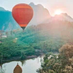 A red and orange hot air balloon floats above green rice fields and a small reflective pond at sunrise, with mountain peaks catching the first light of day—an iconic experience on a 2 week South to North Laos itinerary.