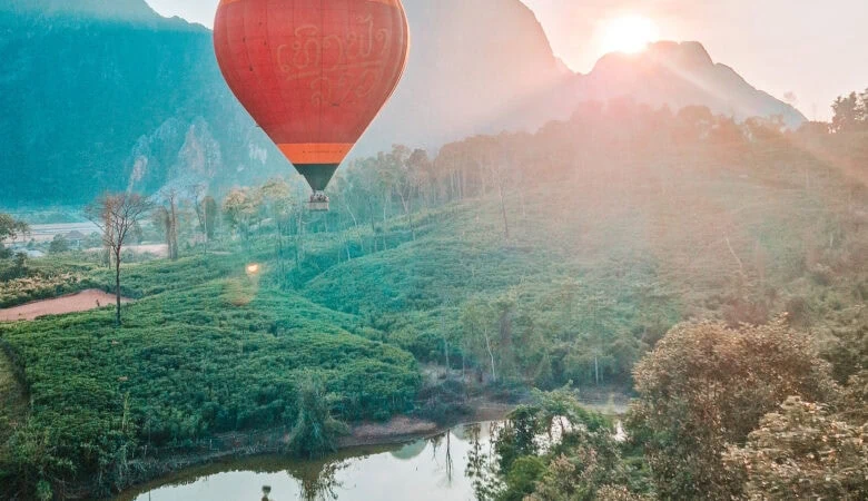 A red and orange hot air balloon floats above green rice fields and a small reflective pond at sunrise, with mountain peaks catching the first light of day—an iconic experience on a 2 week South to North Laos itinerary.