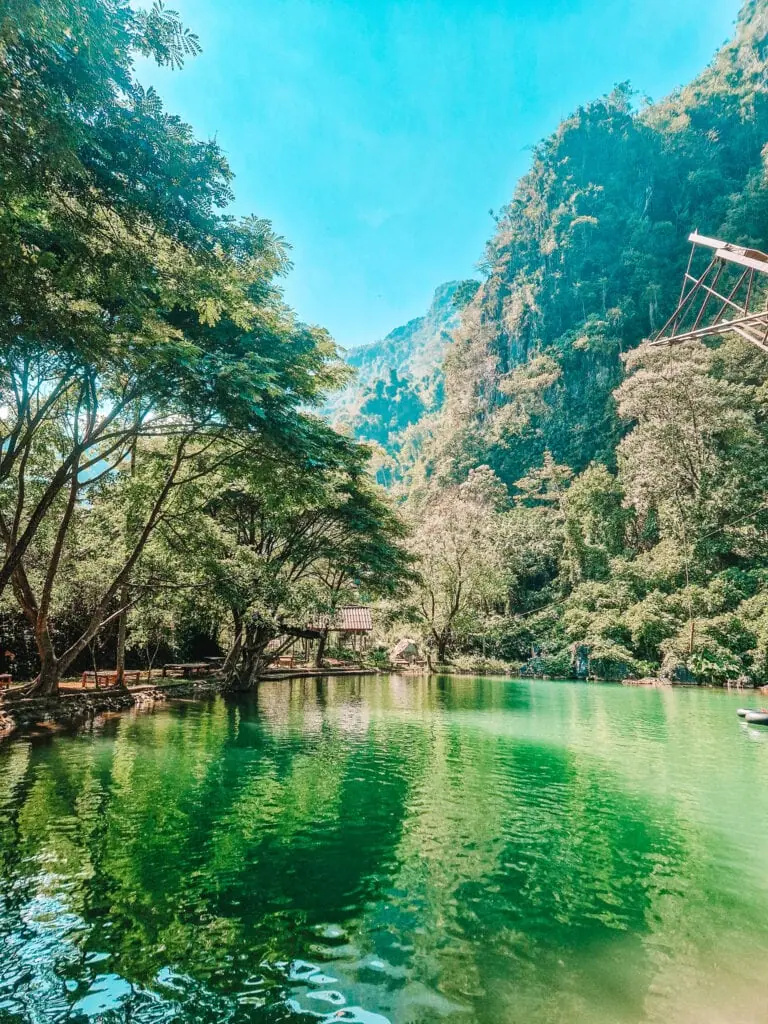 A vibrant turquoise-blue lagoon surrounded by shady trees and towering limestone cliffs, a popular natural swimming spot in Laos.