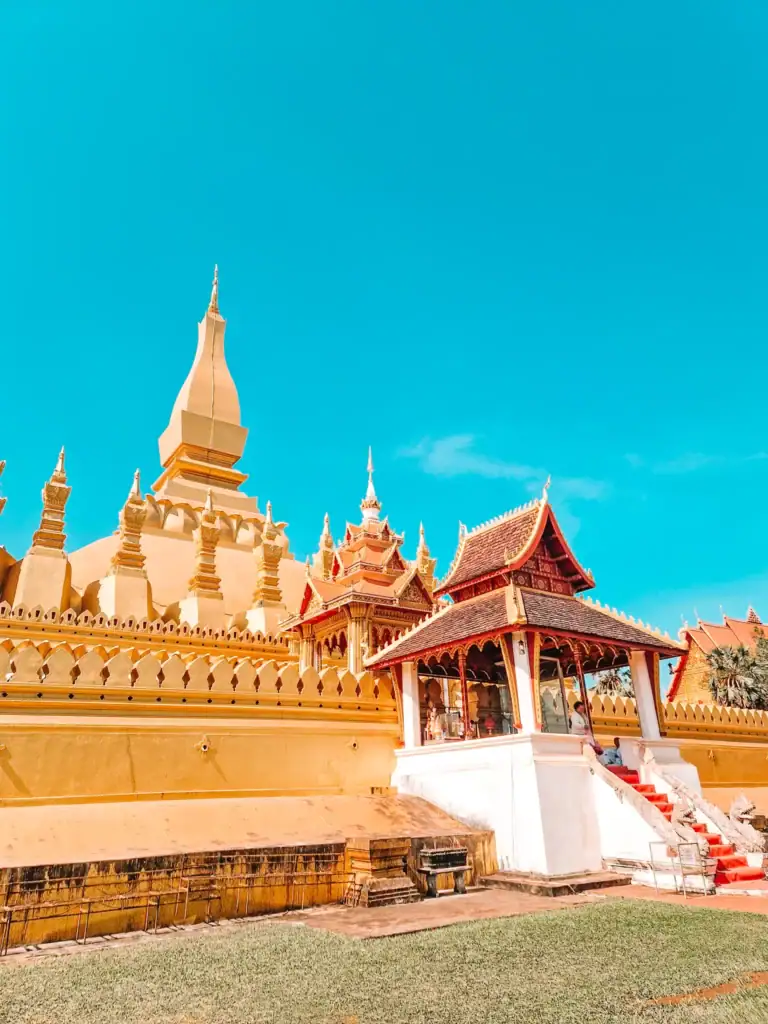 The golden stupa of Pha That Luang shines under a bright blue sky in Vientiane, with its ornate temple structures and manicured grounds.