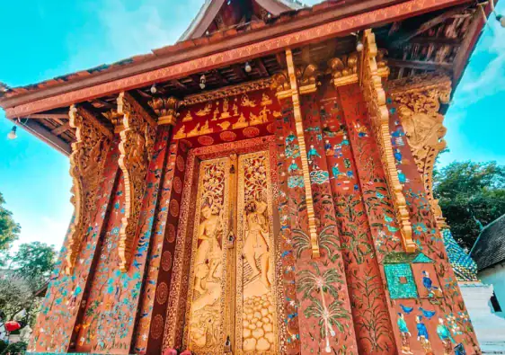 The richly decorated golden doors of Wat Xieng Thong glisten against mosaic-covered walls and blue skies in Luang Prabang. A highlight of temple hopping during a 7 day Laos itinerary.