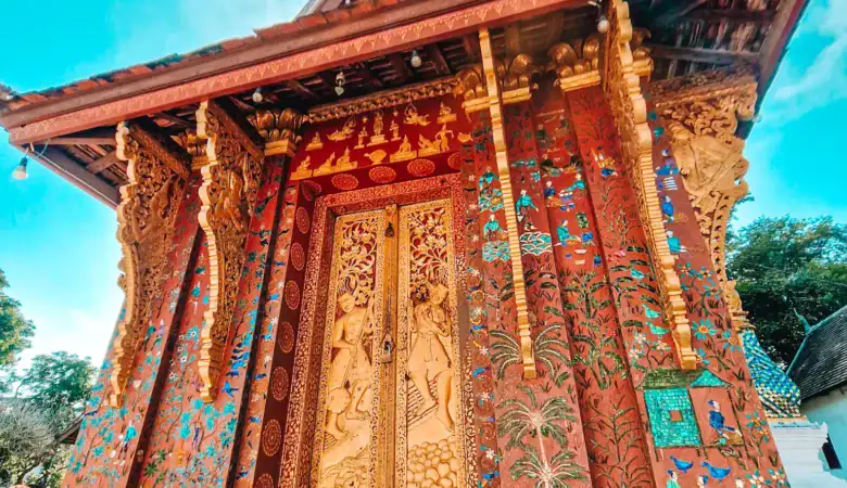 The richly decorated golden doors of Wat Xieng Thong glisten against mosaic-covered walls and blue skies in Luang Prabang. A highlight of temple hopping during a 7 day Laos itinerary.