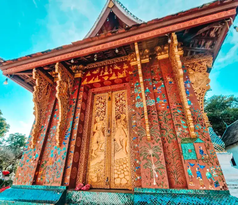 The richly decorated golden doors of Wat Xieng Thong glisten against mosaic-covered walls and blue skies in Luang Prabang. A highlight of temple hopping during a 7 day Laos itinerary.