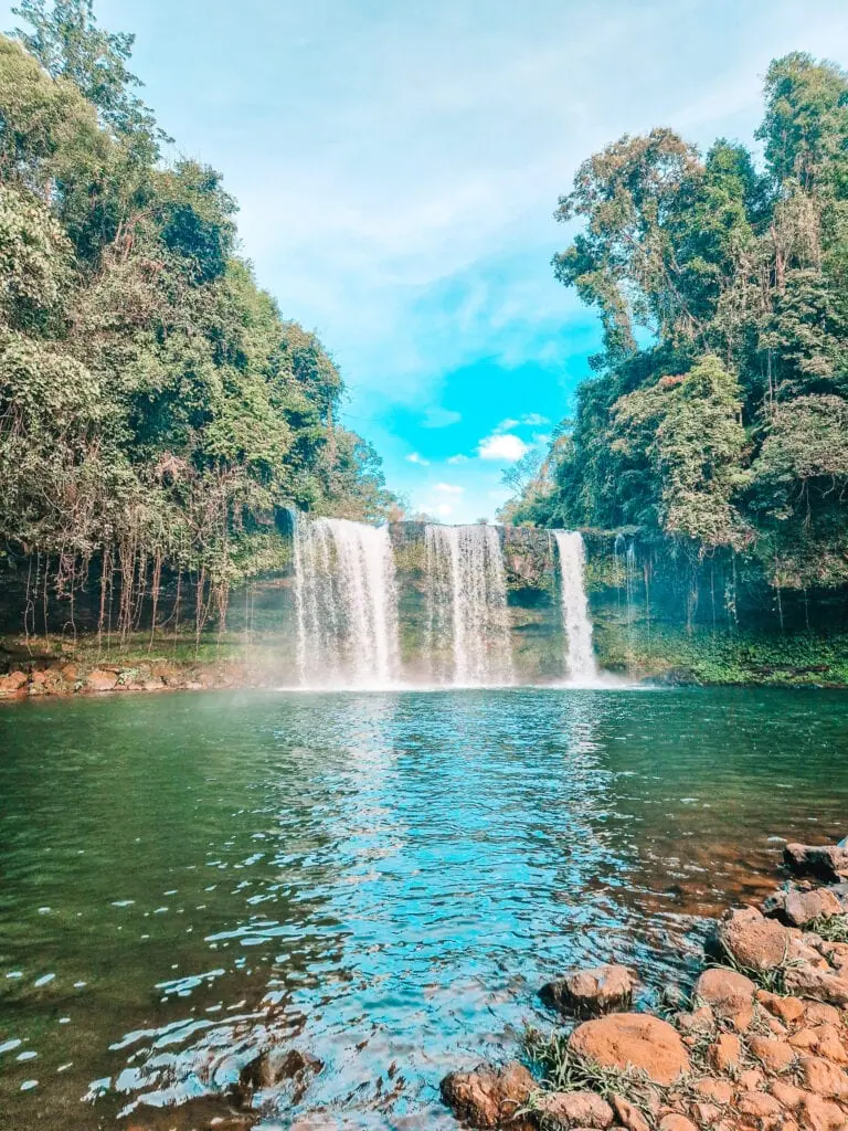 A wide, three-tiered waterfall pours into a clear green pool, framed by lush forest and bright skies in southern Laos.