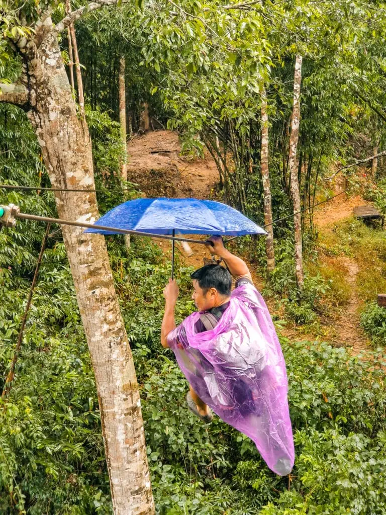A man in a purple poncho and blue umbrella ziplining through light rain, gliding just above the treetops in a misty jungle setting.