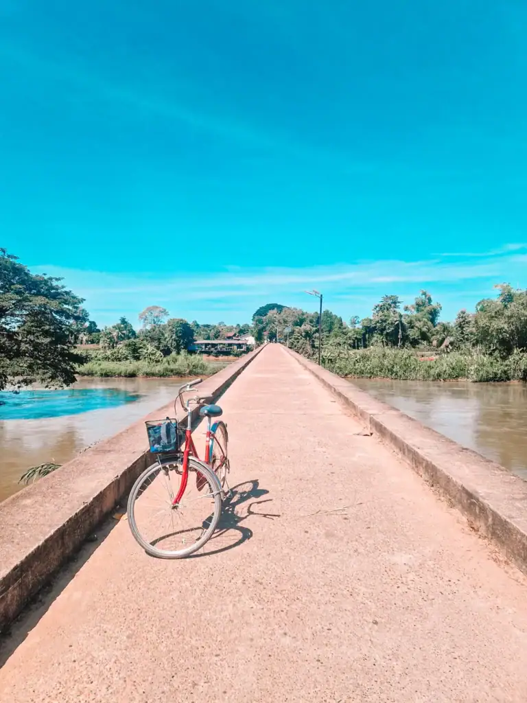 A red bicycle with a front basket leans against the low edge of a narrow concrete bridge stretching across a river under bright blue skies in southern Laos.