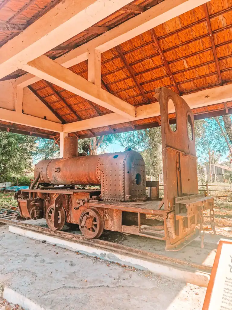 An old, rusted steam engine sits on display under a tiled shelter, part of a historical site in Laos showing remnants of colonial railway infrastructure.