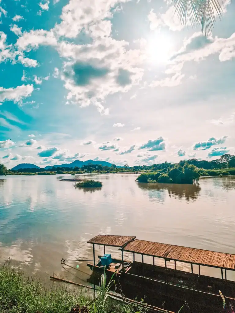 A wooden boat with a thatched roof is moored along the bank of a peaceful river in Laos, with islands, greenery and distant mountains under a glowing sky.
