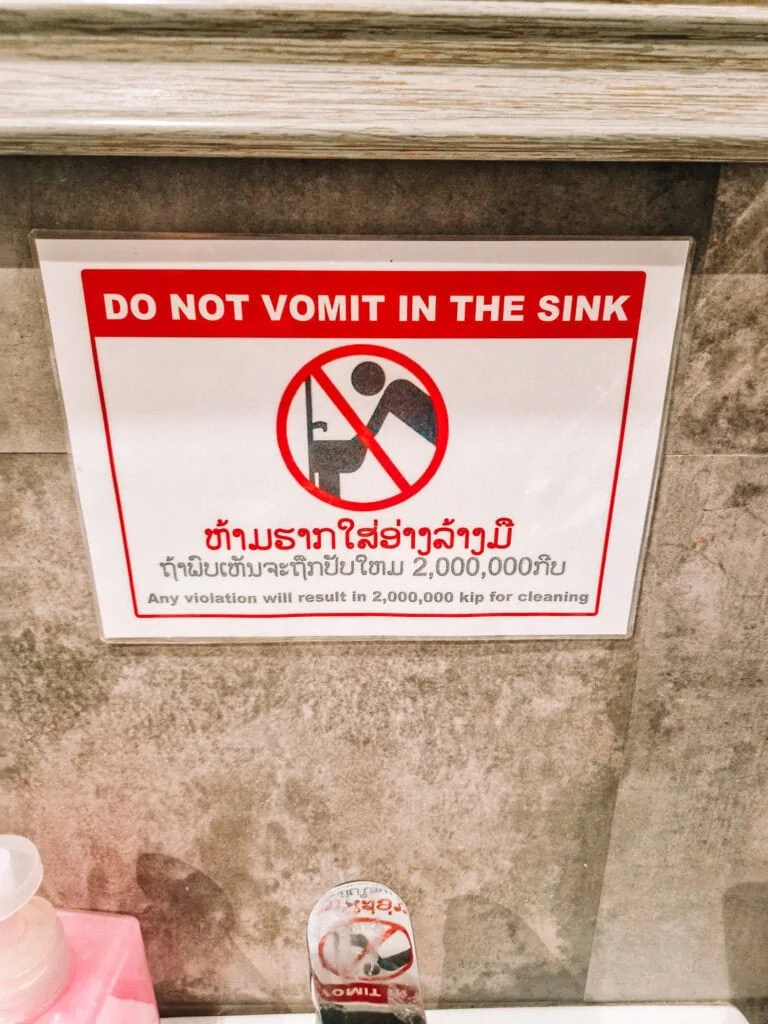 A humorous but stern warning sign above a sink reads “Do not vomit in the sink” in English and Lao, with a fine of 2,000,000 kip for violations.