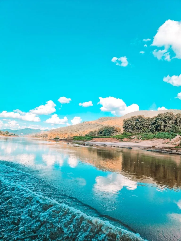 The Mekong River reflects a bright blue sky with fluffy white clouds, as the wake of a boat stirs the otherwise glassy water along a tree-lined shore.