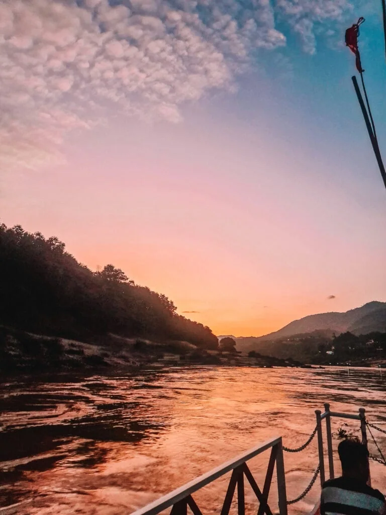 Golden sunset colours reflect on the Mekong River as seen from the side of a slow boat, with silhouetted hills and soft pink clouds overhead. A peaceful river cruise moment on a 2 week north Laos itinerary.