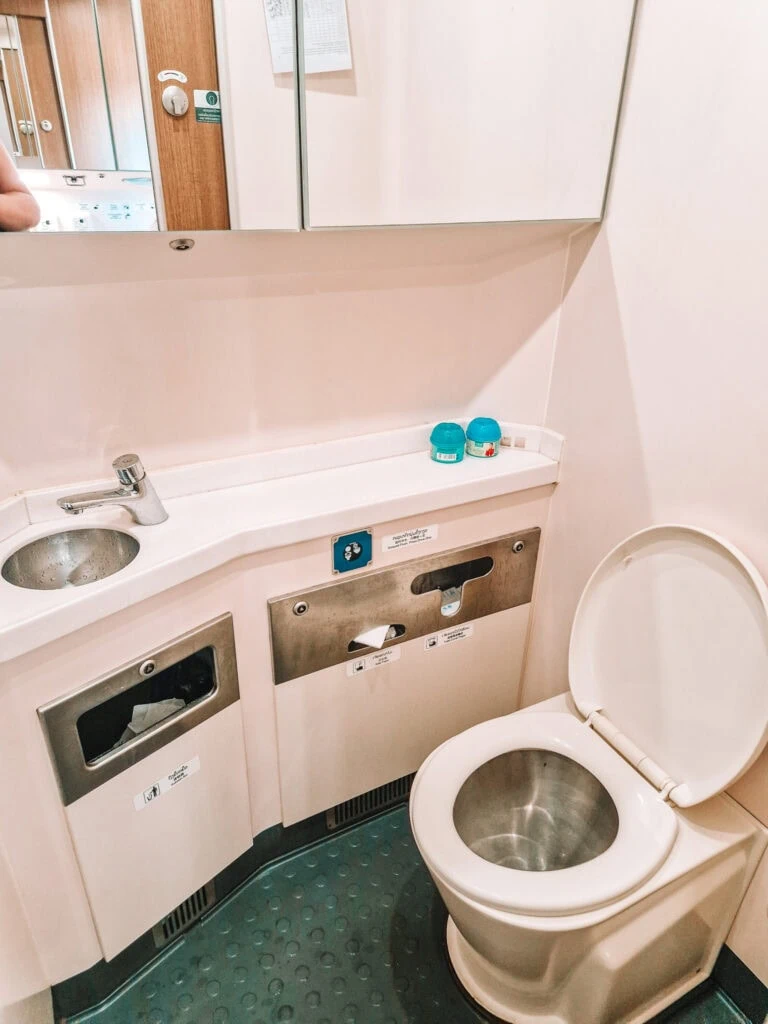 A clean, compact toilet cubicle on the Laos-China Railway with a stainless steel toilet, sink, mirrors and neatly stored toiletries.
