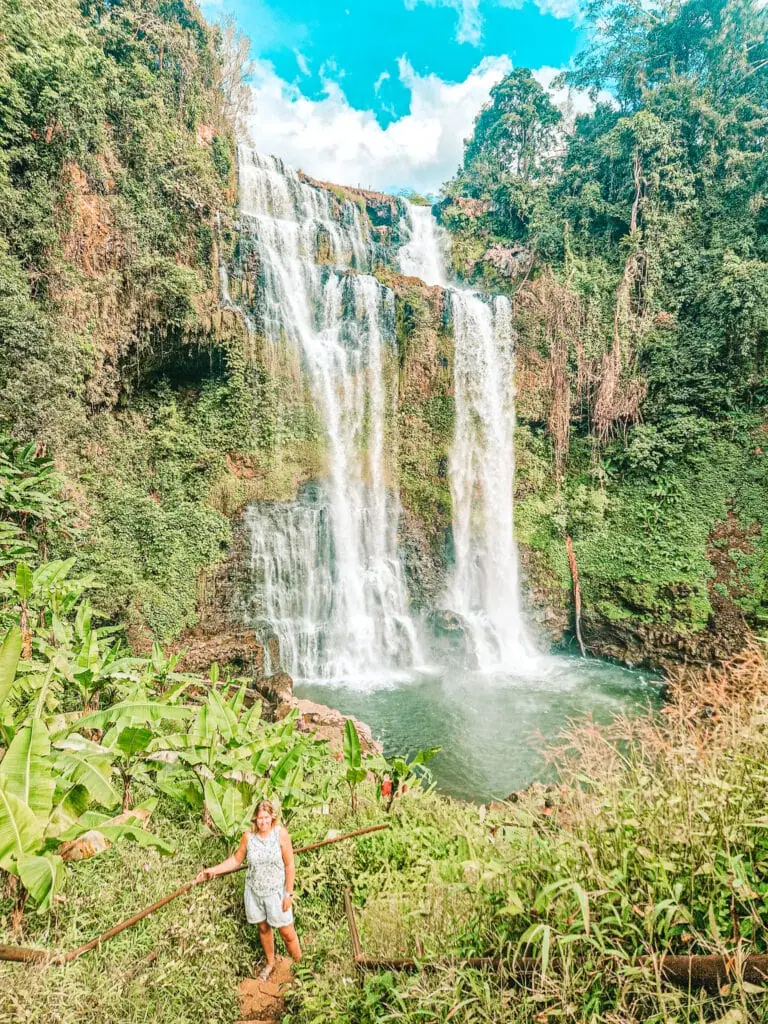 Em stands at the base of a towering jungle waterfall cascading down mossy cliffs, one of the most dramatic natural stops on her 3 week Laos itinerary.