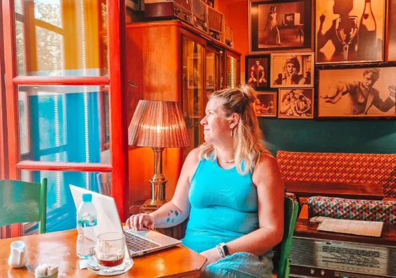 Em sits at a small wooden table working on a laptop inside a cosy vintage style café with warm orange walls, framed photos, and retro decor. This shows the relaxed and characterful café working experience you can expect when choosing between Skopje vs Tirana for digital nomads.