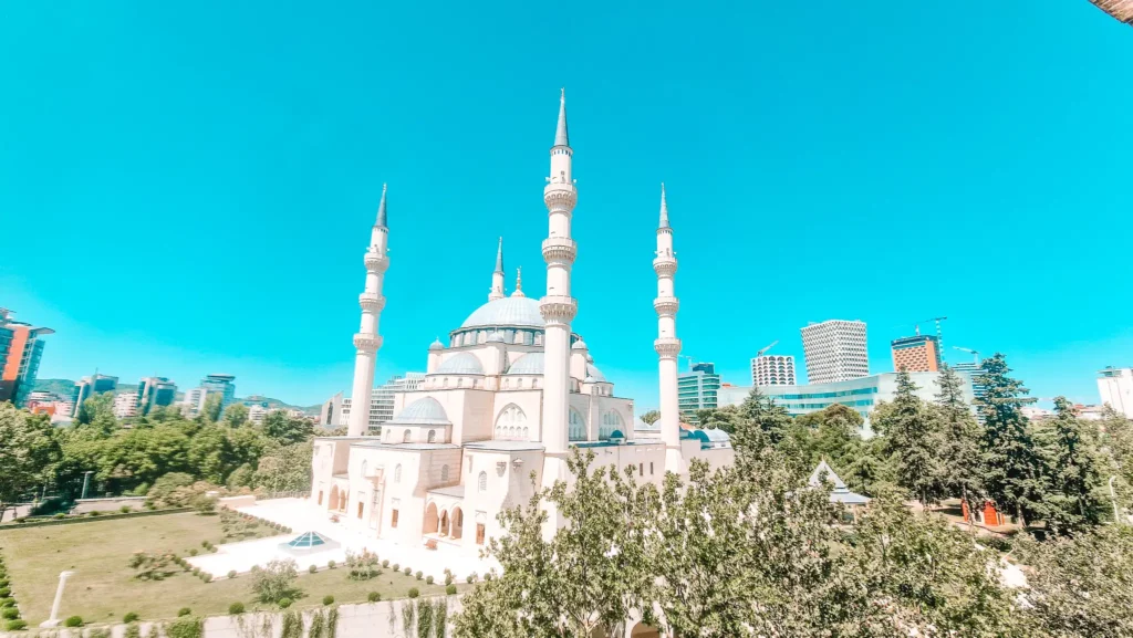 A large white mosque with multiple tall minarets rises above a modern city skyline in Tirana. This shows the mix of traditional and contemporary architecture in daily life.